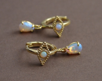 May include: Gold hoop earrings with a teardrop opal dangle and a diamond-shaped charm with a smaller opal in the center. The earrings are on a brown surface.
