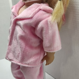 May include: A pink plush doll pajamas set with long sleeves and pants. The pajamas have a button-up front and a collar.