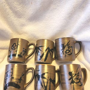 May include: Six brown ceramic mugs with black painted designs. Each mug features a different design, including flowers, bamboo, and Japanese characters. The mugs have a textured finish and curved handles.