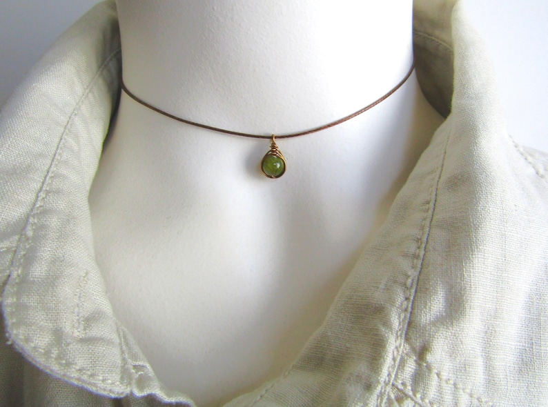 May include: A delicate choker necklace featuring a small, round, green gemstone pendant. The pendant is wire-wrapped in gold-colored metal and hangs from a thin, brown cord. The necklace is displayed on a mannequin wearing a light-colored linen shirt.