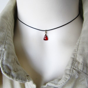 May include: A black choker necklace featuring a small, red, teardrop-shaped pendant. The pendant is a deep red colour and appears to be a faceted gemstone. The necklace is displayed on a white mannequin, with a beige collared shirt in the background.