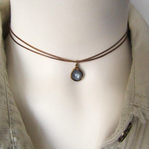 May include: A delicate choker necklace featuring a small, round, pale blue gemstone pendant. The pendant is encased in a gold-colored metal setting and hangs from two thin, brown cords. The necklace is displayed on a white mannequin.