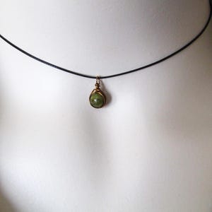 May include: A delicate necklace featuring a small, teardrop-shaped green gemstone pendant. The pendant is encased in a gold-colored wire setting and hangs from a thin, black cord. The necklace is displayed against a neutral background.