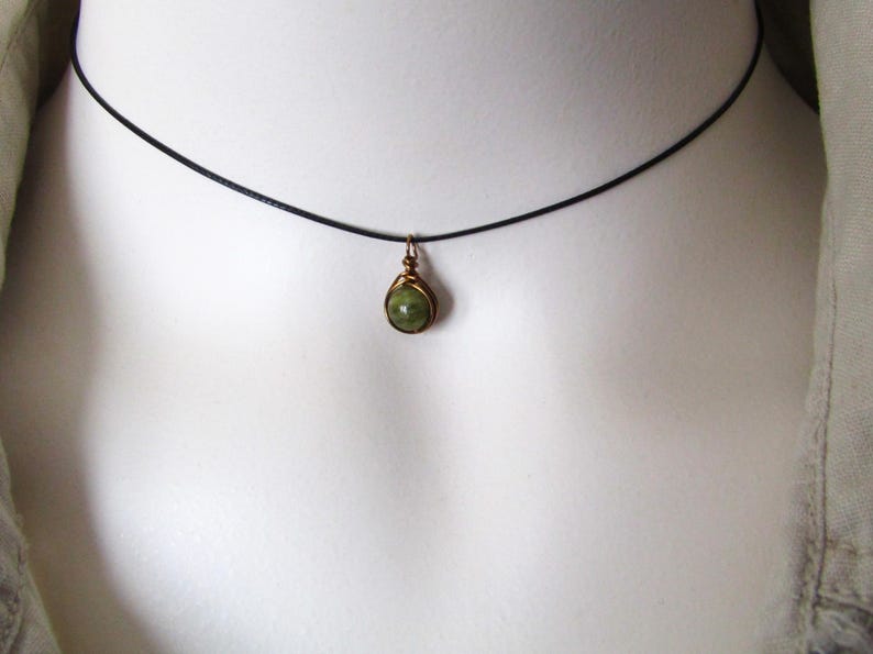 May include: A delicate necklace featuring a small, teardrop-shaped green gemstone pendant. The pendant is encased in a gold-colored wire setting and hangs from a thin, black cord. The necklace is displayed against a neutral background.