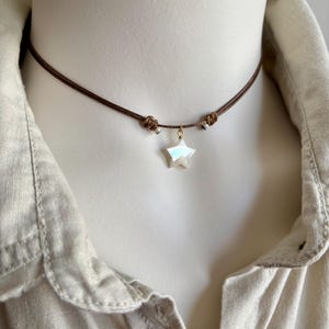May include: A brown cord choker necklace featuring a pearlescent star pendant. The star hangs from a gold-coloured bail. The necklace is knotted on each side and is displayed on a mannequin.