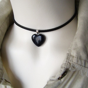 May include: A black choker necklace with a dark blue heart-shaped pendant. The heart pendant has a sparkly, textured appearance. The necklace is displayed on a white mannequin, with a beige collared shirt in the background.