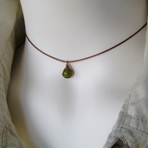 May include: A delicate necklace featuring a small, round, green gemstone pendant. The pendant is encased in a gold-colored wire setting and hangs from a thin, brown cord. The necklace is displayed on a white mannequin.