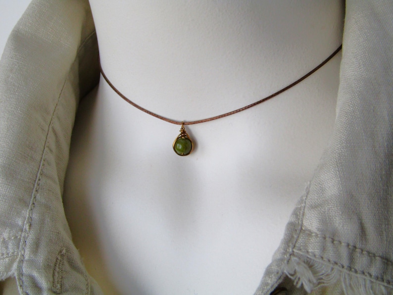 May include: A delicate necklace featuring a small, round, green gemstone pendant. The pendant is encased in a gold-colored wire setting and hangs from a thin, brown cord. The necklace is displayed on a white mannequin.