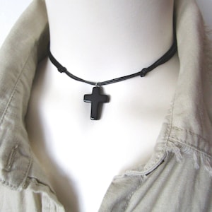 May include: A black cross pendant necklace. The cross is a solid, dark color and hangs from a black cord. The necklace is displayed on a white mannequin, with a beige collared shirt in the background.