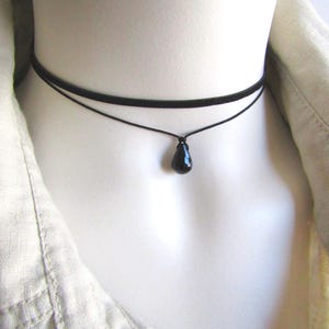 May include: A black choker necklace with a teardrop pendant. The necklace has two strands, one thicker and one thinner, with a small, shiny black pendant hanging from the bottom strand. The necklace is displayed on a mannequin.