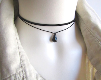May include: A black choker necklace with a teardrop pendant. The necklace has two strands, one thicker and one thinner, with a small, shiny black pendant hanging from the bottom strand. The necklace is displayed on a mannequin.