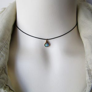 May include: A black choker necklace with a teardrop-shaped pendant. The pendant has a blue and green iridescent stone, set in gold-coloured metal. The necklace is displayed on a white mannequin.