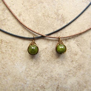 May include: Two necklaces with green gemstone pendants. Each pendant is a round, green stone wrapped in gold-colored wire. The necklaces have black and brown cords. The background is a light brown, textured surface.