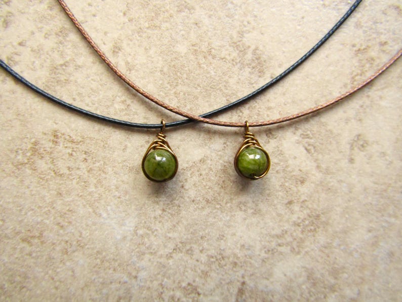 May include: Two necklaces with green gemstone pendants. Each pendant is a round, green stone wrapped in gold-colored wire. The necklaces have black and brown cords. The background is a light brown, textured surface.