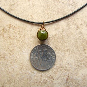 May include: A handmade pendant necklace featuring a green gemstone wrapped in copper wire. The pendant hangs from a black cord. A dime is placed below the pendant for size comparison. The necklace is displayed on a neutral-toned surface.