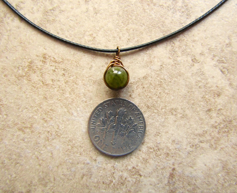 May include: A handmade pendant necklace featuring a green gemstone wrapped in copper wire. The pendant hangs from a black cord. A dime is placed below the pendant for size comparison. The necklace is displayed on a neutral-toned surface.