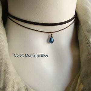 May include: A two-strand choker necklace with a Montana blue teardrop pendant. The top strand is a dark brown suede-like material, and the bottom strand is a thin brown cord. The pendant is a faceted blue crystal. The text "Color: Montana Blue" is visible.