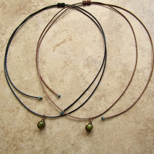 May include: Three delicate necklaces with thin cords in black, brown, and blue. Each necklace features a small, olive-green gemstone pendant. The necklaces are displayed on a neutral, textured surface.