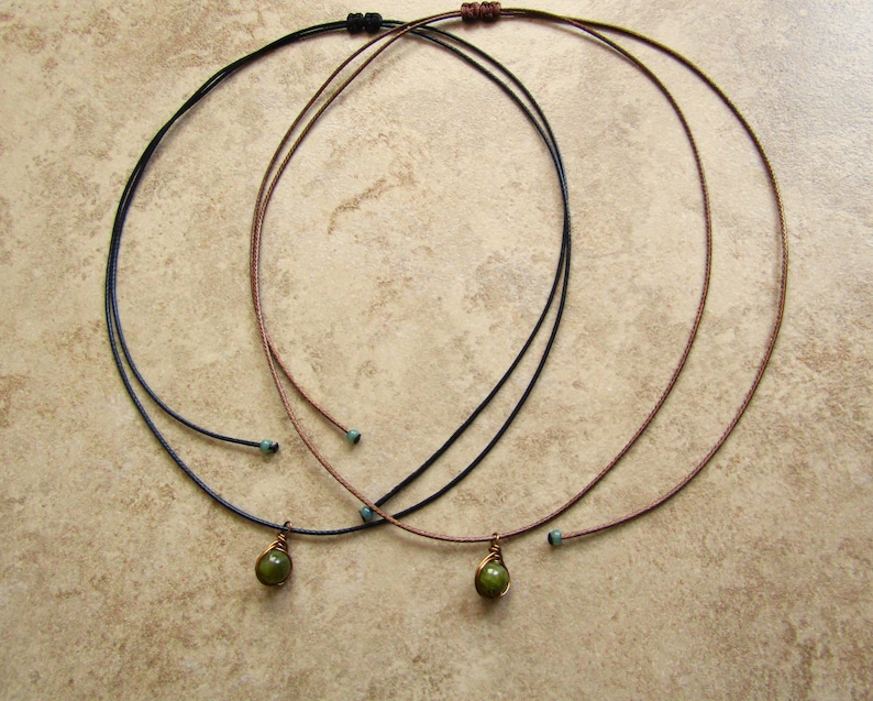 May include: Three delicate necklaces with thin cords in black, brown, and blue. Each necklace features a small, olive-green gemstone pendant. The necklaces are displayed on a neutral, textured surface.