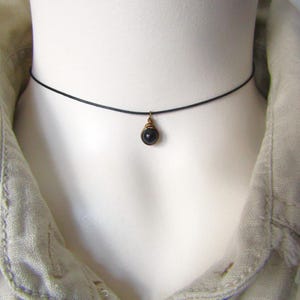 Peut inclure: Un collier ras du cou noir avec un petit pendentif en pierre précieuse sombre. Le pendentif est enroulé dans un fil de métal doré. Le collier est présenté sur un mannequin blanc, avec une chemise beige en arrière-plan.