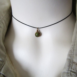 May include: A delicate choker necklace featuring a small, green gemstone pendant. The pendant is wire-wrapped in a gold-toned metal and hangs from a thin, black cord. The necklace is displayed on a white mannequin, with a light green jacket in the background.