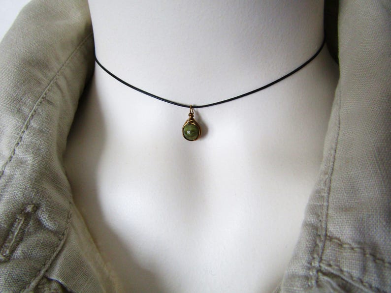 May include: A delicate choker necklace featuring a small, green gemstone pendant. The pendant is wire-wrapped in a gold-toned metal and hangs from a thin, black cord. The necklace is displayed on a white mannequin, with a light green jacket in the background.