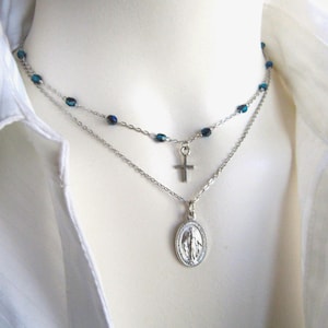 May include: A layered necklace featuring a silver chain with a cross pendant and a pendant with a religious image. The top layer has blue beads. The necklace is displayed against a white shirt.