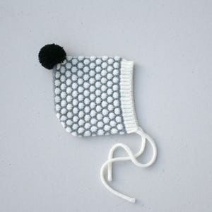 May include: A white and grey knitted baby bonnet with a black pom-pom on top. The bonnet has a white tie.
