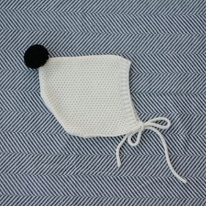 May include: A white knitted baby bonnet with a black pom-pom on top. The bonnet has a tie at the back.