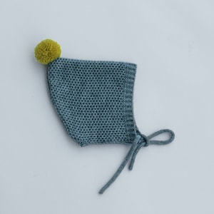 Puede incluir: Un gorro de bebé de punto azul con un pompón amarillo en la parte superior. El gorro tiene un lazo en la parte posterior.