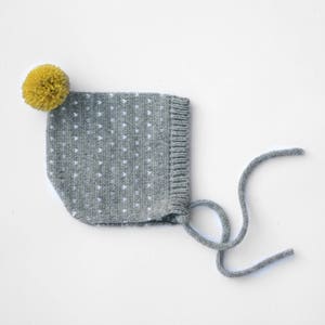 Può includere: Un berretto per neonato di lana grigia con pois bianchi e un pon pon giallo in cima.