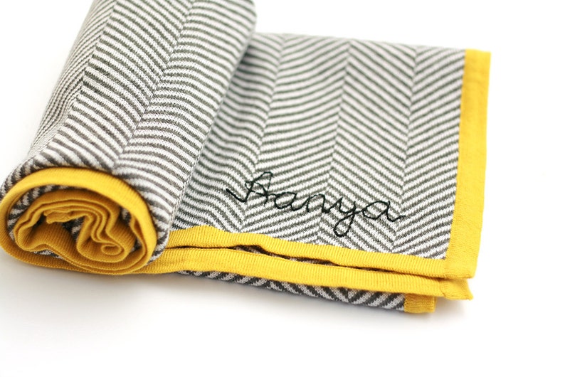Puede incluir: Una manta de beb&eacute; con estampado de chevron gris y blanco con un borde amarillo y el nombre "Banya" bordado en hilo negro.