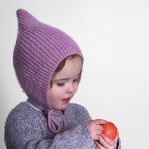 Pode incluir: Uma criança pequena usando um gorro de tricô roxo com uma longa ponta pontiaguda. A criança está segurando uma bola vermelha em suas mãos.