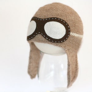 May include: A brown knitted hat with a white stripe and a brown felt aviator goggles design. The hat has ear flaps.
