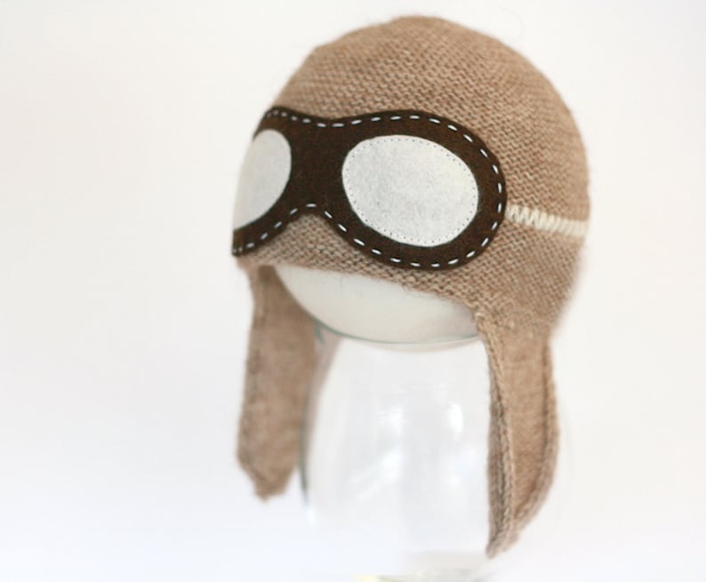 May include: A brown knitted hat with a white stripe and a brown felt aviator goggles design. The hat has ear flaps.