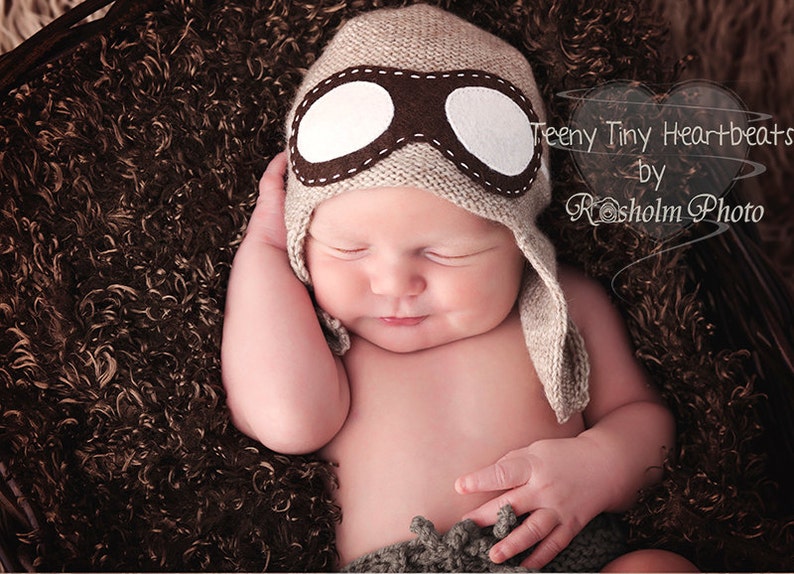 May include: A newborn baby wearing a brown and white aviator hat with a brown fur trim, sleeping on a brown fuzzy blanket. The text "Teeny Tiny Heartbeats by Rosholm Photo" is in the upper right corner of the image.