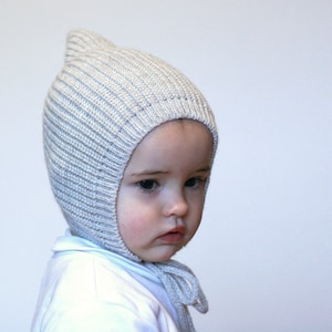 May include: A baby wearing a light grey knitted hooded hat with a pointed top. The hat has a ribbed texture and covers the baby's ears.