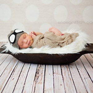 May include: A newborn baby wearing a gray and white aviator hat sleeps in a brown wooden bowl lined with white fur. The baby is wrapped in a beige blanket.