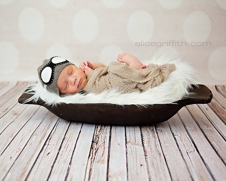 May include: A newborn baby wearing a gray and white aviator hat sleeps in a brown wooden bowl lined with white fur. The baby is wrapped in a beige blanket.