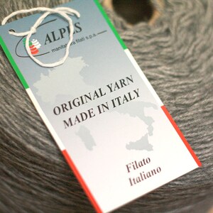 May include: A white tag with a green and red border, featuring the text "ALPS" and "ORIGINAL YARN MADE IN ITALY". The tag is attached to a gray ball of yarn.