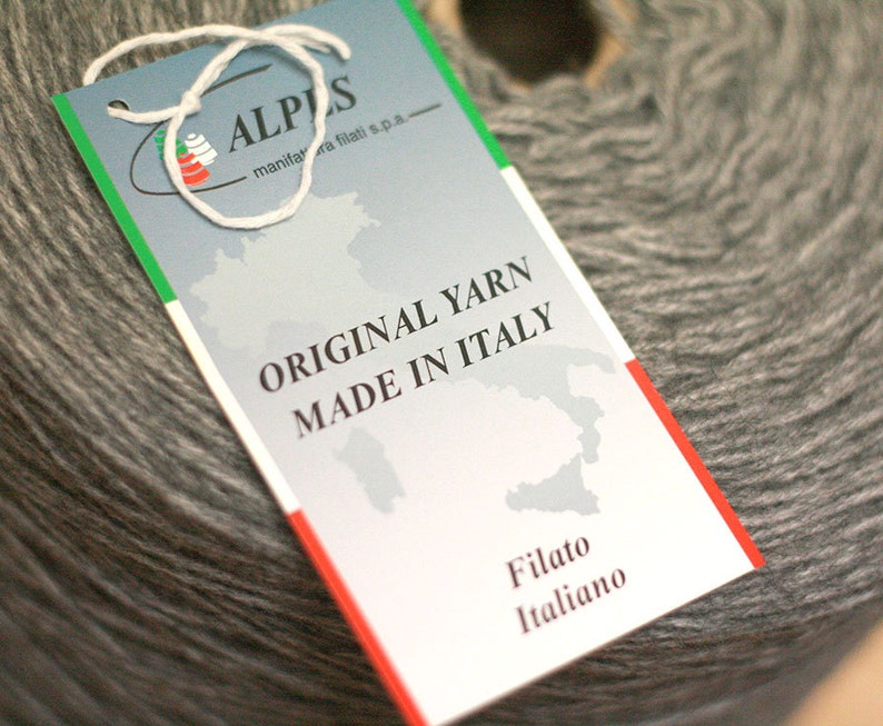 May include: A white tag with a green and red border, featuring the text "ALPS" and "ORIGINAL YARN MADE IN ITALY". The tag is attached to a gray ball of yarn.