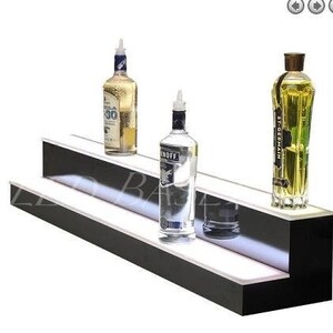 May include: Two-tiered black shelf with a white LED light strip. The shelf is displaying three glass bottles of liquor. The bottles are clear glass with labels. The top shelf has a bottle of St. Germain elderflower liqueur and the bottom shelf has a bottle of Stoli vodka and a bottle of tequila.