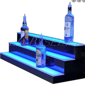 May include: Three-tiered black shelf with blue LED lights, designed to hold bottles of liquor. The top shelf has a bottle of vodka, the middle shelf has a bottle of tequila, and the bottom shelf has a bottle of rum.