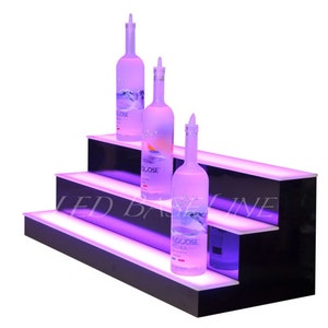 May include: Three-tiered black acrylic liquor bottle display with LED lights. The display has three shelves with a purple glow. Three clear glass bottles of vodka are on the shelves. The bottles are labeled "Goose Vodka".
