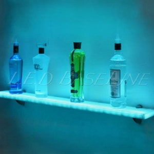 May include: A glowing, illuminated shelf displays four bottles of liquor against a blue backdrop. The bottles vary in shape and color, with clear, green, and blue liquids. The shelf is lit with a soft, cool light, creating a modern, stylish display.