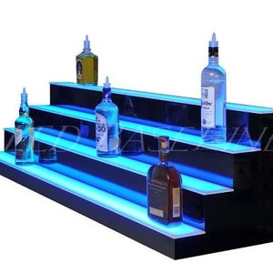 May include: A black, three-tiered liquor display shelf with blue LED lighting. Several bottles of liquor are arranged on the shelves. The display is designed to showcase bottles in a bar or home setting.