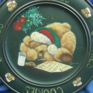 May include: A green ceramic plate with the word "Cookies" written around the edge in gold lettering. A teddy bear wearing a Santa hat is sleeping on a bed of cookies with a glass of milk. The plate is decorated with a Christmas tree and ornaments.