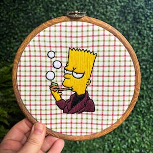 May include: Embroidered hoop art featuring a yellow cartoon character with blue eyes and a red jacket, blowing bubbles. The character is holding a pipe in their mouth. The background is a red and green checkered fabric.