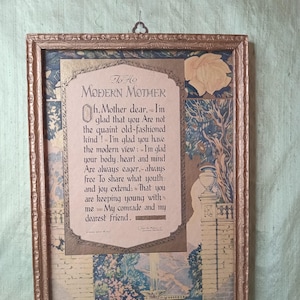 May include: A framed print with a rose-gold colored frame. The print features a poem titled "To a Modern Mother" with decorative floral and landscape illustrations in shades of blue, beige, and gold. The print is hung by a decorative metal hook.