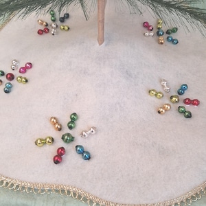 May include: A white felt Christmas tree skirt featuring a gold scalloped trim along the edge. The surface is decorated with clusters of small, shiny, multi-colored glass bead ornaments in red, green, blue, gold, and silver arranged in floral patterns.